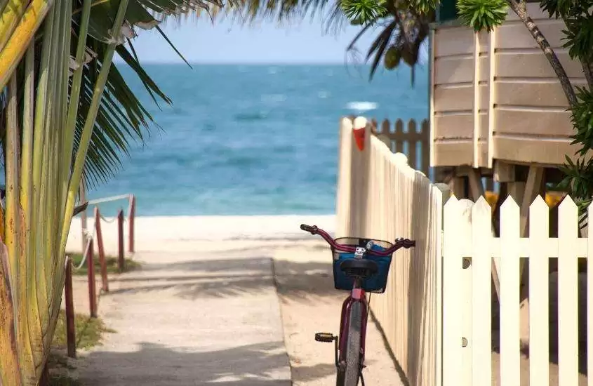 durante o dia, bicicleta rosa, com cestinha, encostada em cerca branca de casa e, ao fundo, mar