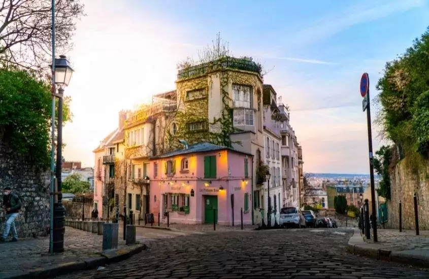 durante o dia, pessoas caminhando em frente a construção rosa, em rua de pedras de Montmartre, um lugar onde ficar em paris