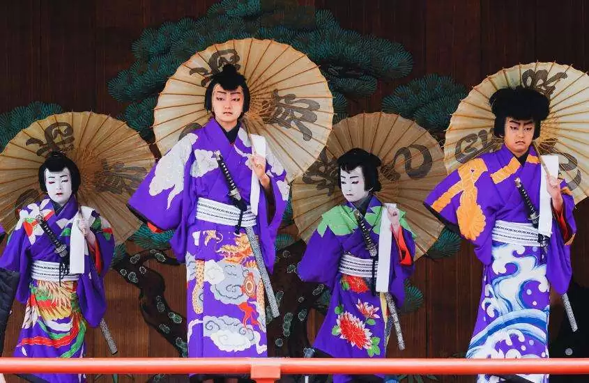 atores, maquiados e vestidos com quimonos azuis, segurando guarda-chuva com escritos em japonês, se apresentam em teatro tradicional japonês