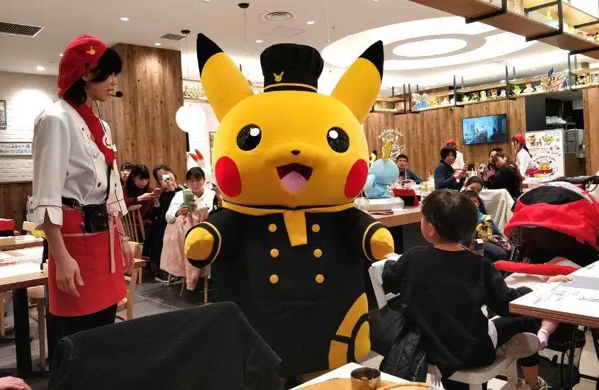 pessoas, em mesas de restaurante, observam pessoa fantasiada de Pikachu