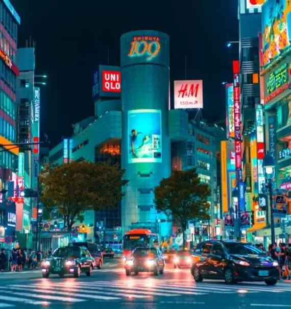 durante a noite, carros e pessoas passando em rua de um bairro de tokyo, cheio de prédios com letreiros iluminados