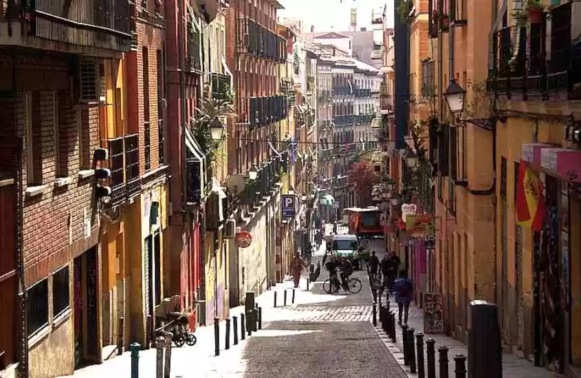 durante o dia, pessoas caminhando e pedalando em rua inclinada, cercada de prédios em lavapiés, um lugar onde ficar em Madri