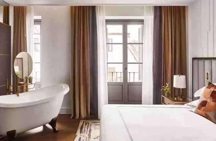durante o dia, banheira branca, espelho redondo e cama de casal em suíte de hotel 5 estrelas em madri com varanda
