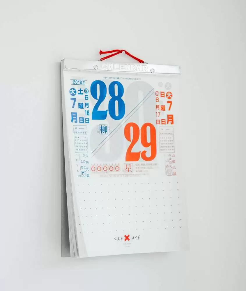 calendário branco, com números azuis e laranjas e palavras em japonês, pendurado em parede branca