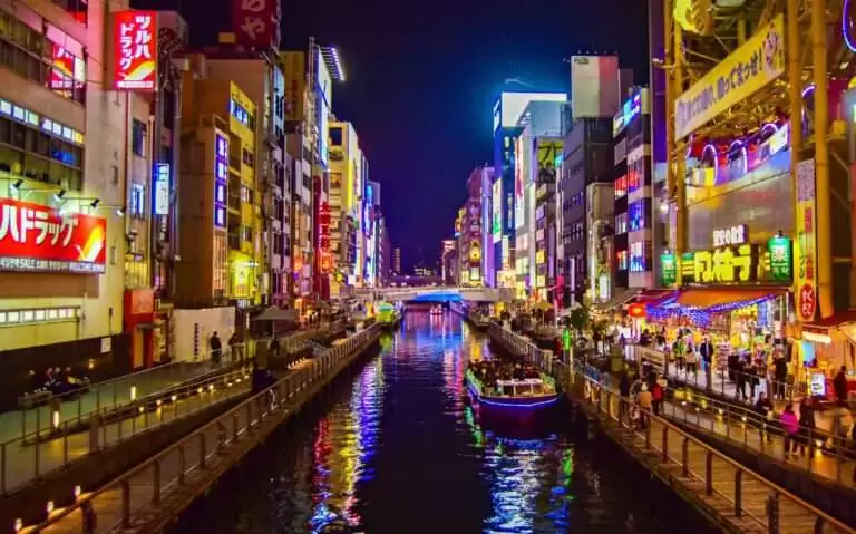 durante a noite, pessoas em barco iluminado passando em rio cercado de prédios com letreiros coloridos e frases em japonês em osaka japão