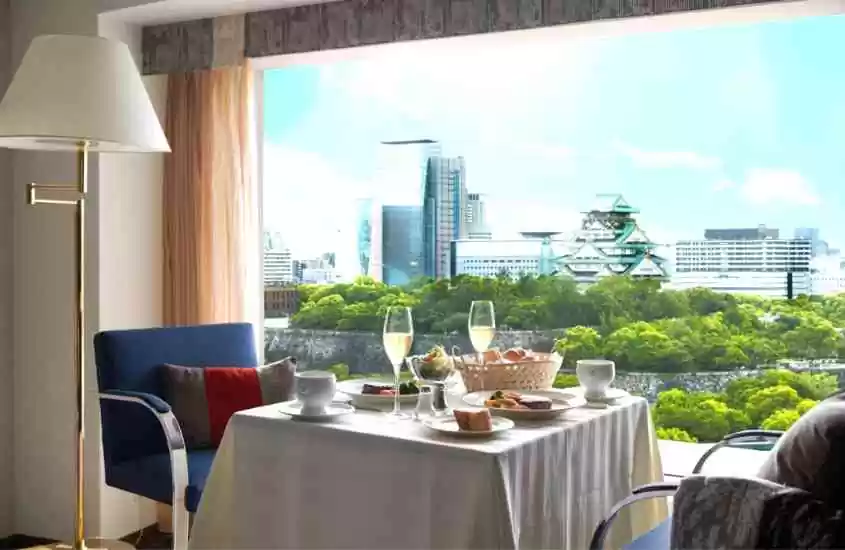 durante o dia, abajur, cadeiras ao redor de mesa, onde há taças de champanhe e pratos de comida, em suíte de hotel com vista para o castelo de osaka