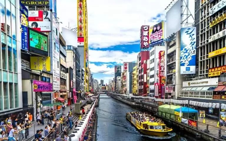 durante o dia, pessoas em barco amarelo passando em rio cercado de prédios com letreiros coloridos e frases em japonês em osaka japão
