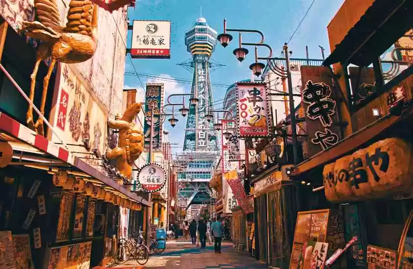 durante o dia, pessoas caminhando em beco rodeado por lojas com letreiros coloridos em japonês