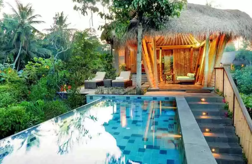 durante o dia, bangalô de palha em frente a piscina retangular cercada de árvores em resort onde ficar em bali