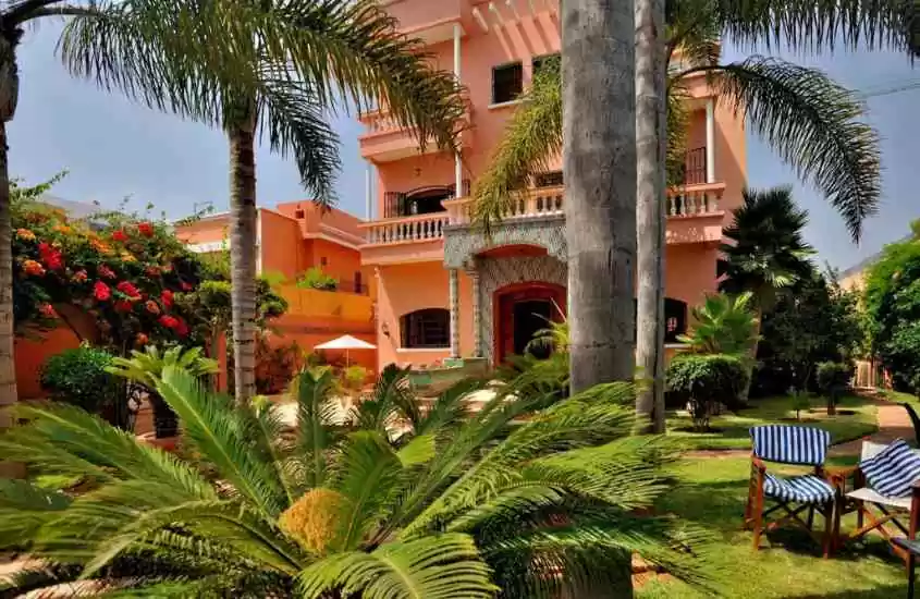 durante dia ensolarado, cadeiras em quintal cheio de árvores, em frente a casa rosa onde funciona um hotel em Casablanca