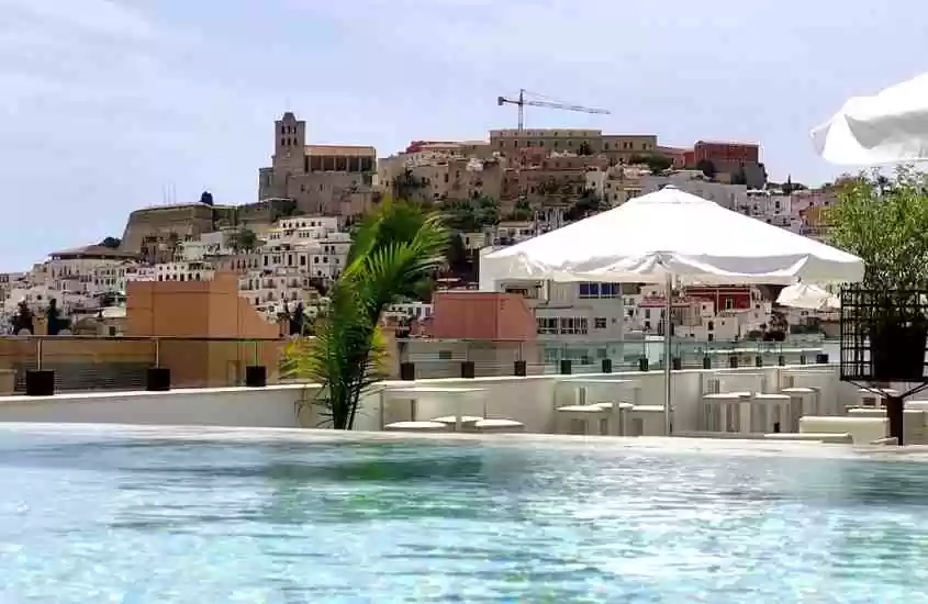 durante o dia, guarda-sol, mesas e cadeiras e brancas em frente a piscina em cobertura de hotel em ibiza com vista para a cidade