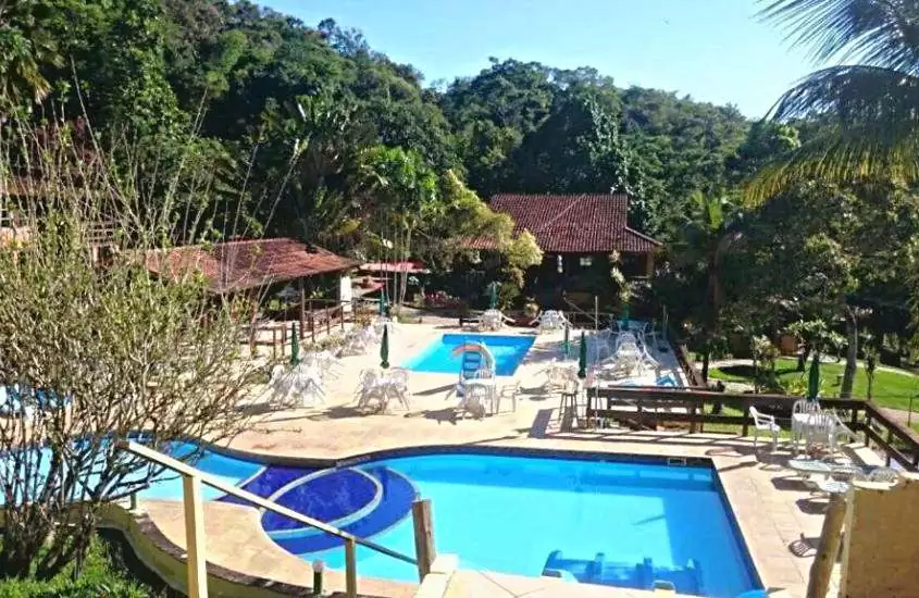 durante dia ensolarado, piscinas ao ar livre e cadeiras e mesas brancas em área de lazer de hotel rodeada de árvores