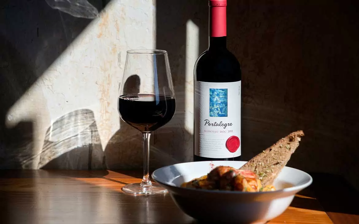 taça de vinho tinto, garrafa de vinho Portalegre e um sanduíche sobre um prato branco, dispostos em cima de uma mesa de madeira, iluminados pela luz do sol