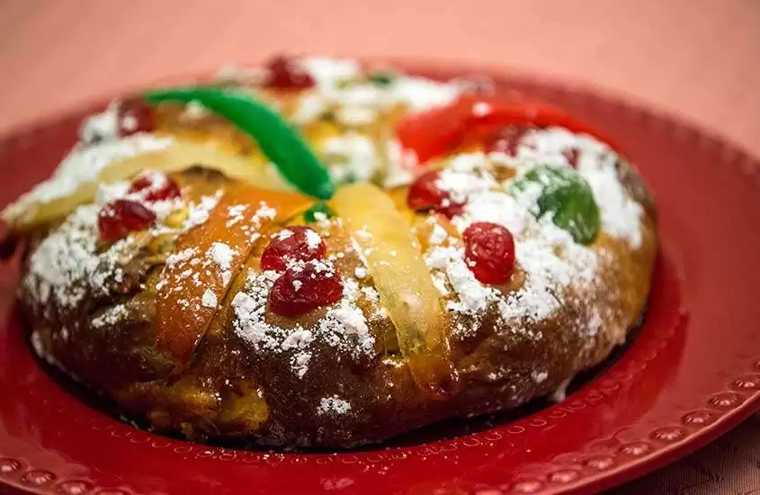 Bolo Rei, uma sobremesa de Portugal, servido em um prato redondo vermelho com frutas cristalizadas, nozes e açúcar de confeiteiro por cima