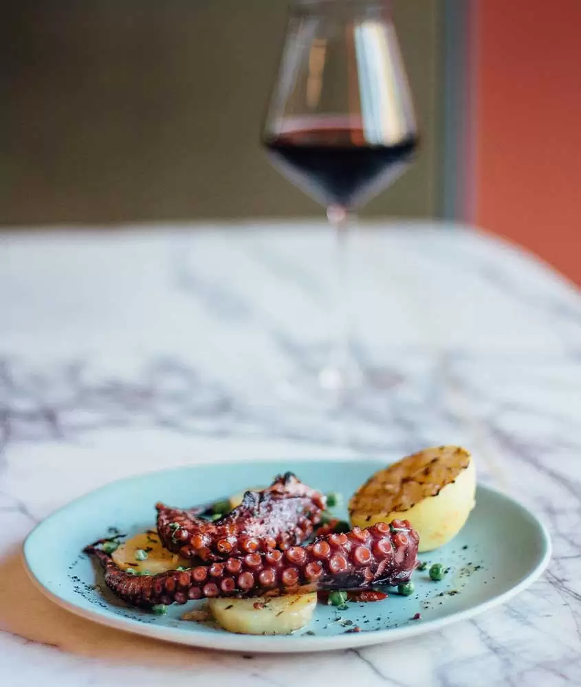 Polvo à lagareiro, um prato da culinária portuguesa, servido em um prato branco redondo e acompanhado por uma taça de vinho tinto, sobre uma mesa branca