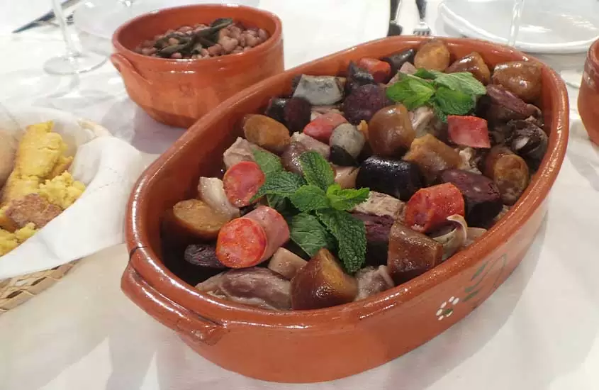 Uma travessa oval de barro com cozido à portuguesa, contendo carne, legumes e batatas, disposto em camadas e servido em cima de uma mesa branca.