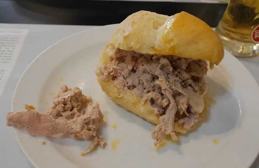 Sanduíche de bifana da culinária portuguesa, com pão branco, carne de porco temperada e molho, servido em prato branco redondo sobre uma mesa.