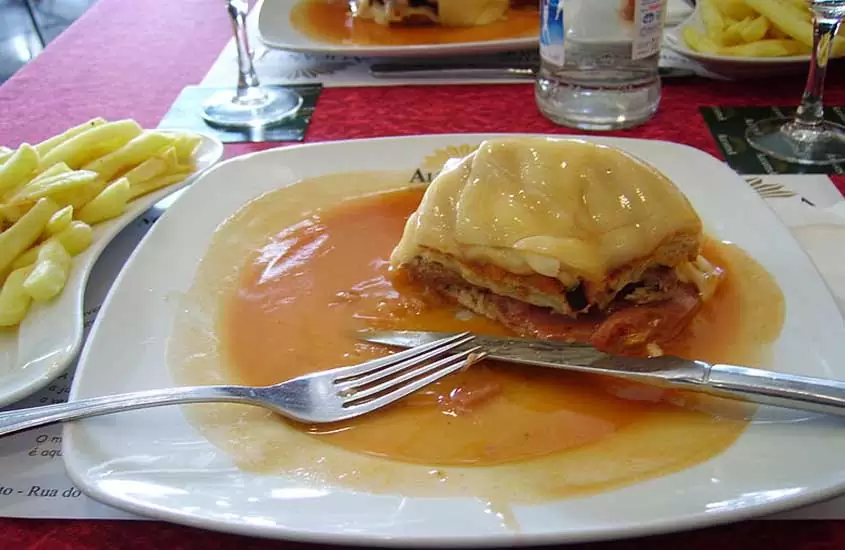 garfo, faca e pedaço de francesinha, sanduíche da culinária portuguesa, em cima de prato branco quadrado