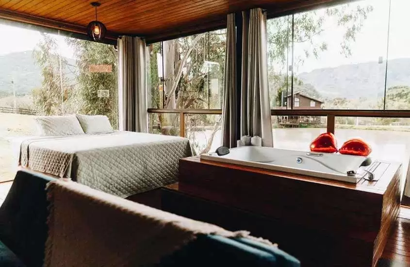 durante o dia, cama de casal e banheira em chalé com grande janelas de vidro com vista para a natureza de Urubici SC