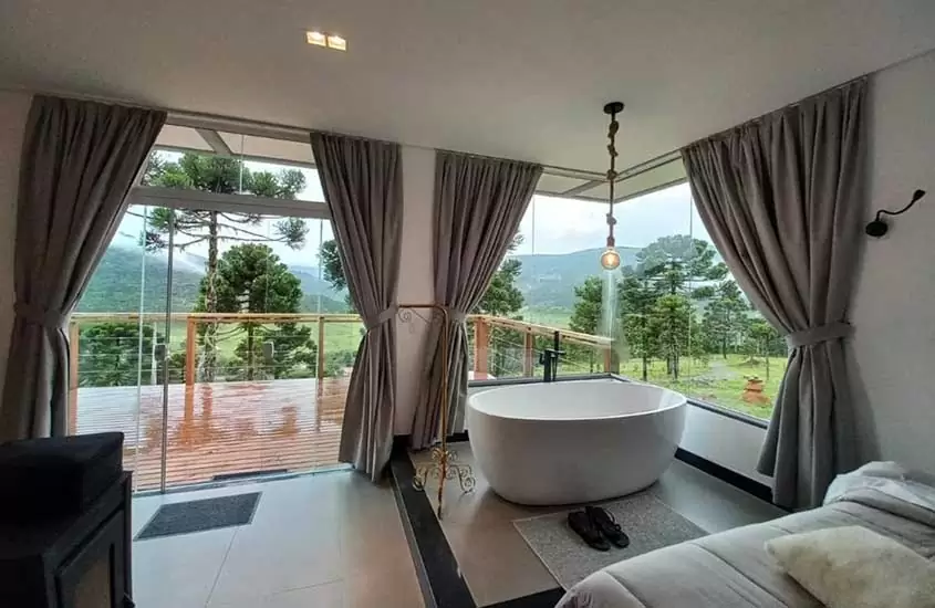 durante o dia, cama de casal, lareira e banheira em chalé com amplas janelas de vidro com vista para a natureza de Urubici SC, em dia nublado