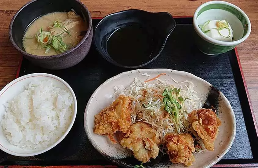 Foto de cima de um teishoku, refeição japonesa em bandeja preta com várias seções para diferentes pratos, incluindo arroz, salada, tempura, sashimi e molhos, servido em uma mesa de madeira