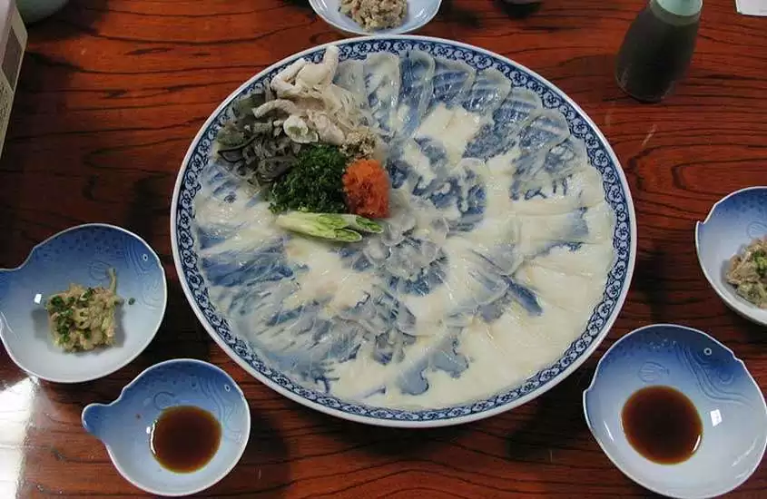 prato branco decorado com desenhos azuis, contendo fatias de fugu (peixe baiacu), um prato típico da culinária japonesa