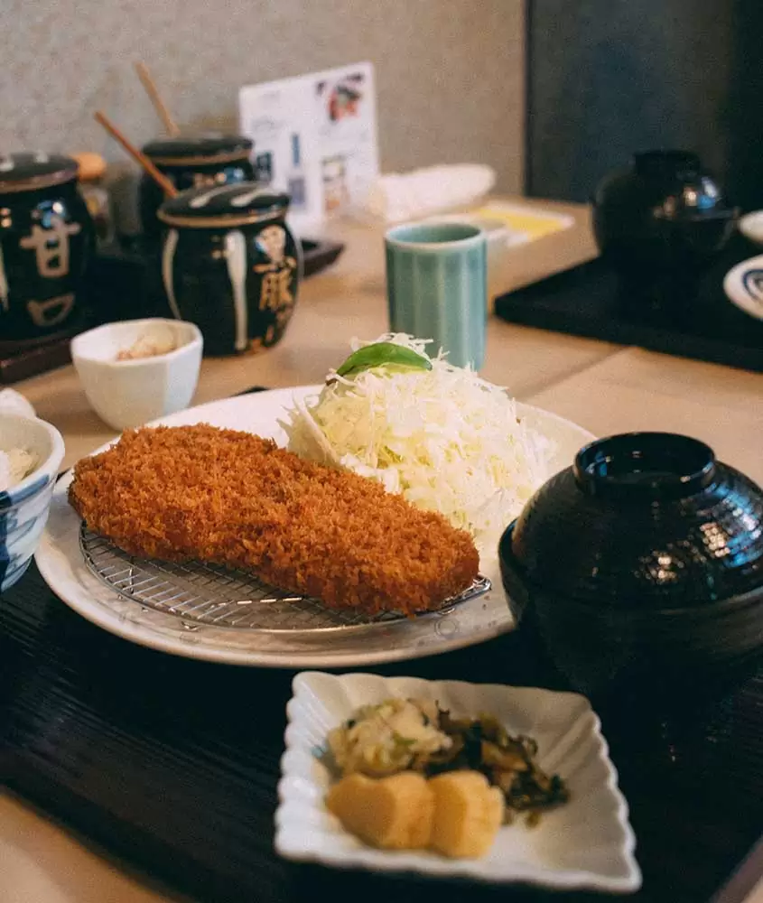 Tonkatsu, um prato japonês com carne de porco empanada e frita, servido em um prato branco redondo com um molho à parte e guarnecido com legumes