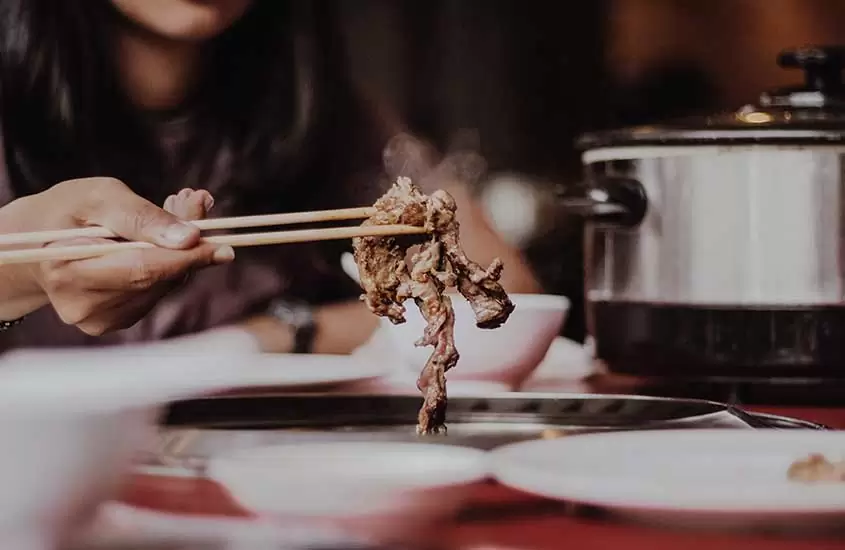 Pessoa segurando com hashi um pedaço de yakiniku grelhado, servido em um prato branco sobre a mesa de madeira
