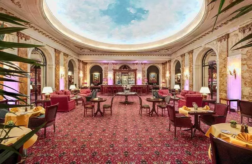 Salão sofisticado de hotel em Bruxelas, com sofás vermelhos, cadeiras e mesas de madeira, e um teto com pintura que simula o céu