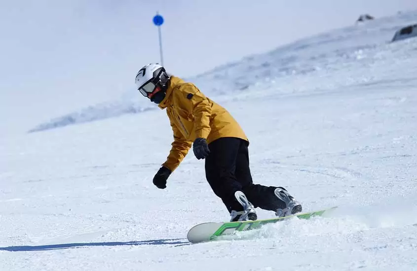 Durante o dia ensolarado, um praticante de snowboard com capacete branco, óculos de proteção, casaco amarelo, calça preta e luvas pretas, em ação em uma pista de esqui em Bariloche durante o inverno