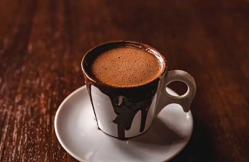 Chocolate quente cremoso servido em uma xícara de porcelana branca, sobre uma mesa de madeira rústica
