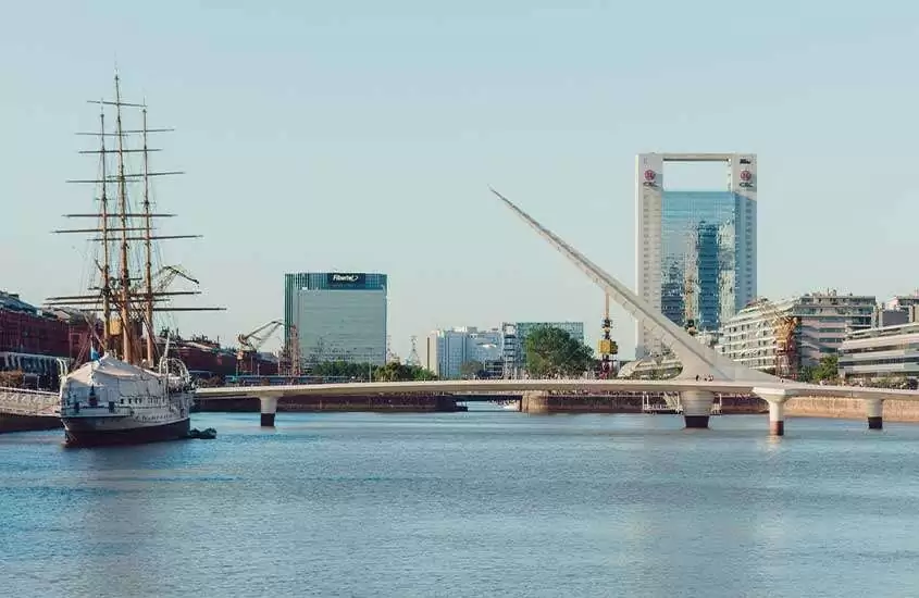 Durante o dia, barco passando pelo rio e, ao fundo, possível a ponte Puente de la Mujer e prédios modernos em Puerto Madero, um dos bairros para se hospedar em Buenos Aires