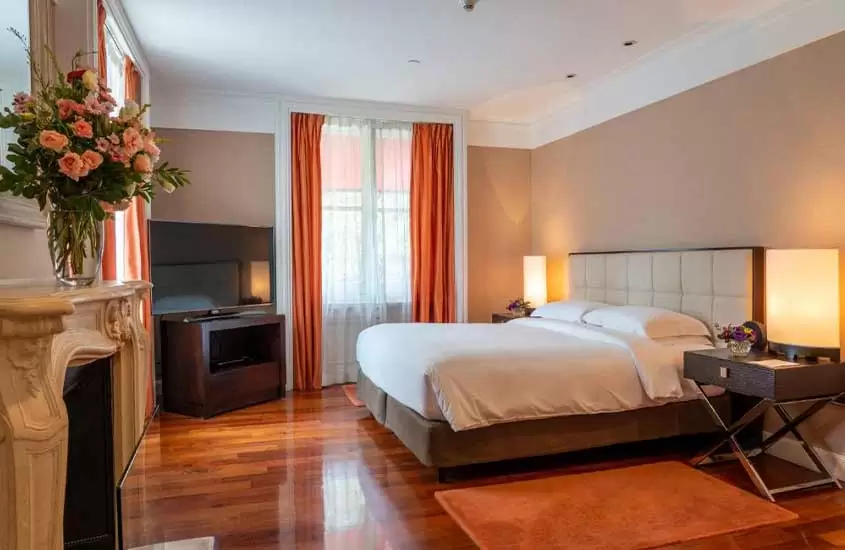 Durante o dia, suíte de hotel em Buenos Aires, com piso de madeira, cama de casal e uma lareira com um vaso de flores rosas em cima