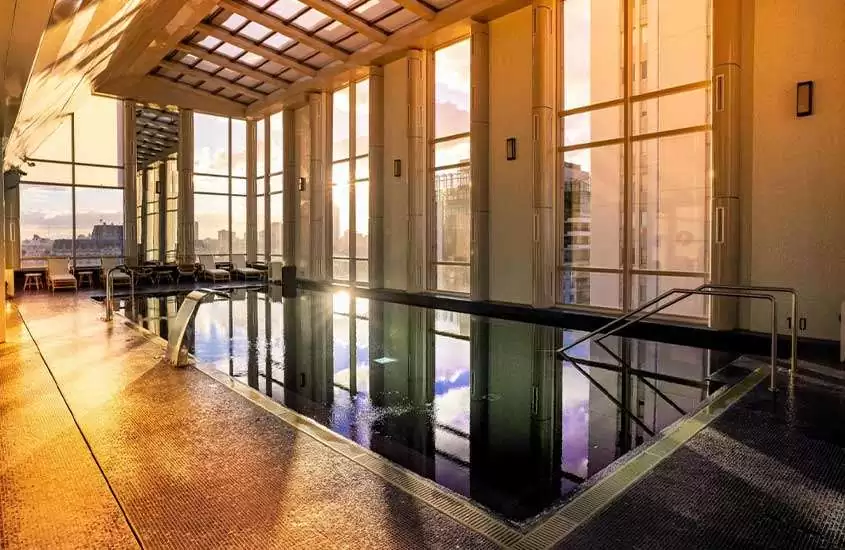 Durante um entardecer, uma piscina retangular iluminada em uma área de lazer coberta de um hotel. Através das grandes janelas panorâmicas, é possível ver a cidade de Buenos Aires ao fundo