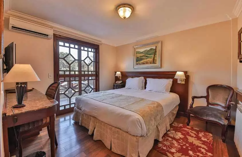 cama de casal e poltrona de madeira acolchoada em suíte de hotel em campos do jordão com piso de madeira, varanda e vista para a cidade