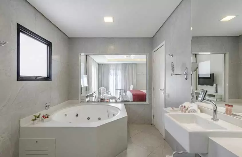 banheira de hidromassagem e pia de mármore, em banheiro com ampla janela com vista para cama de casal em quarto de hotel para passar o réveillon em campos do jordão