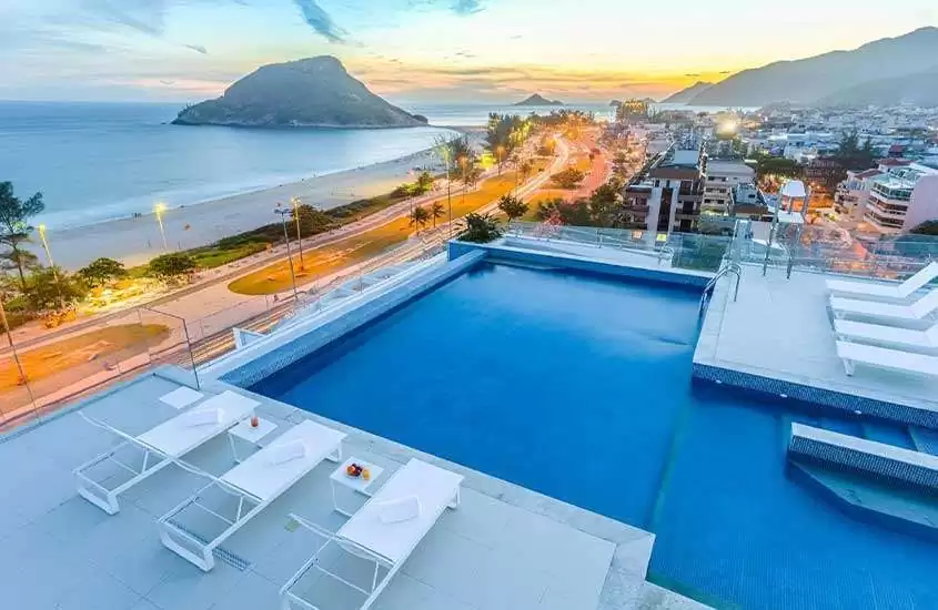 durante entardecer, espreguiçadeiras e piscina em cobertura de hotel no recreio dos bandeirantes com vista para o mar.