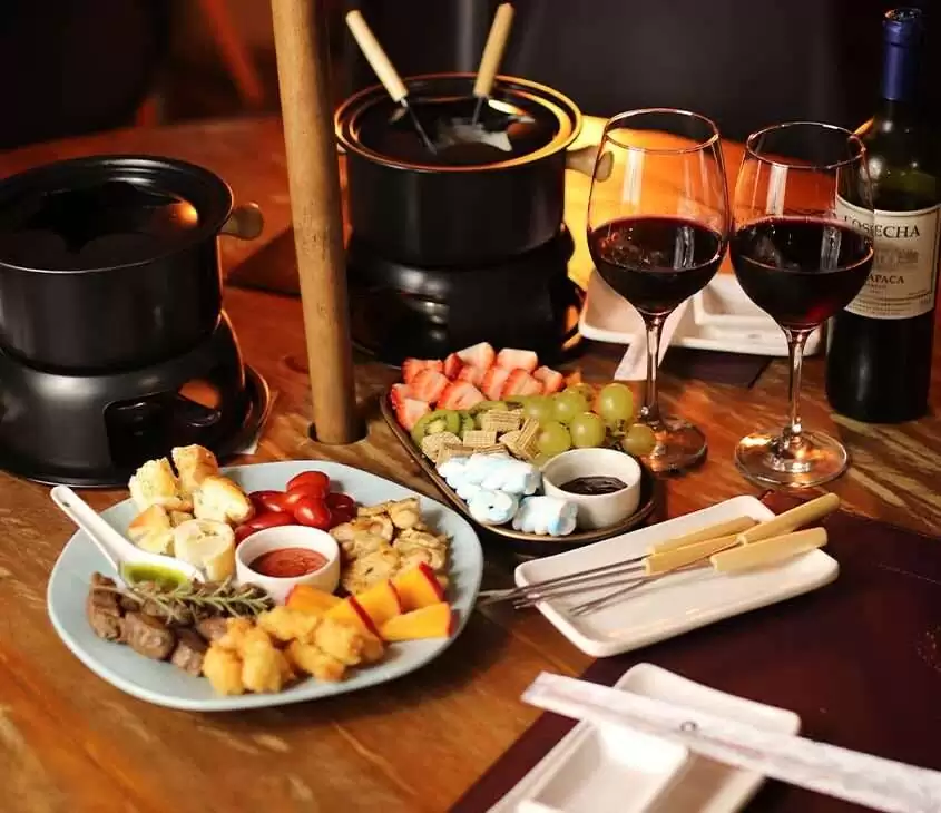 panelas de fondue, uma seleção de frios e petiscos variados, dispostos em tábuas e travessas e taças de vinho em mesa de madeira de um restaurante em bariloche