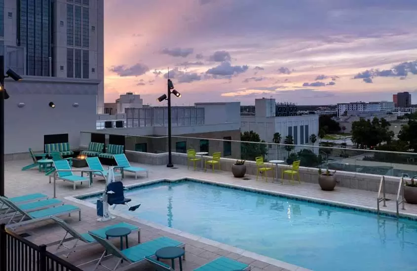 durante entardecer, vista aérea de espreguiçadeiras azuis em frente a piscina retangular em cobertura de hotel em orlando com vista para a cidade