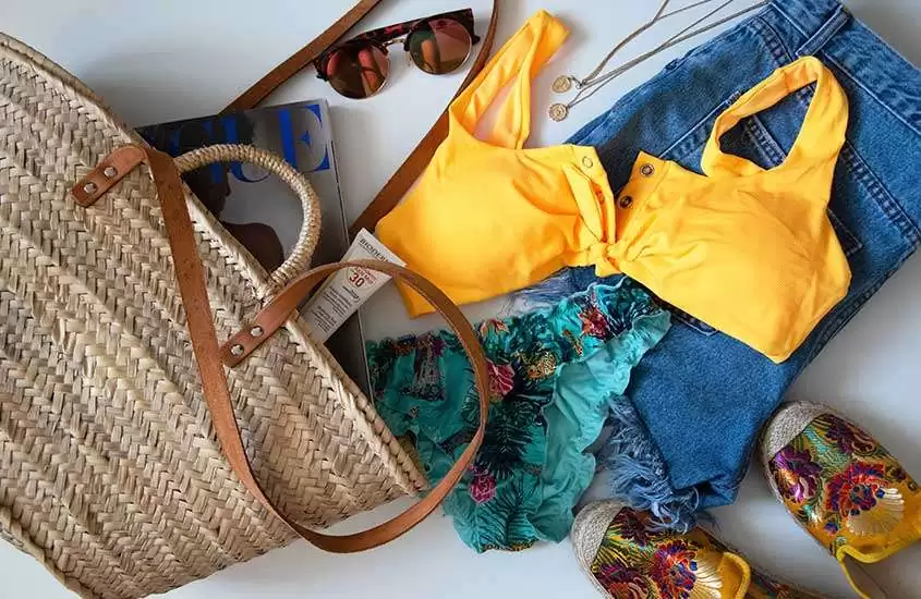 bolsa de palha, protetor solar, revista, óculos de sol, blusa amarela, short jeans, biquíni azul e sapatilhas em cima de mesa branca