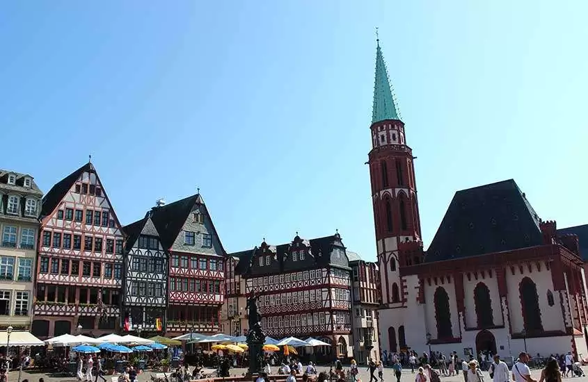 durante um dia ensolarado, pessoas caminhando em praça com igreja e casas em estilo enxaimel em Römerberg, um dos pontos turísticos em Frankfurt