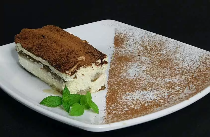 pedaço de tiramisu, doce italiano feito com biscoitos embebidos em café, creme de mascarpone e cacau em pó, servido em prato quadrado
