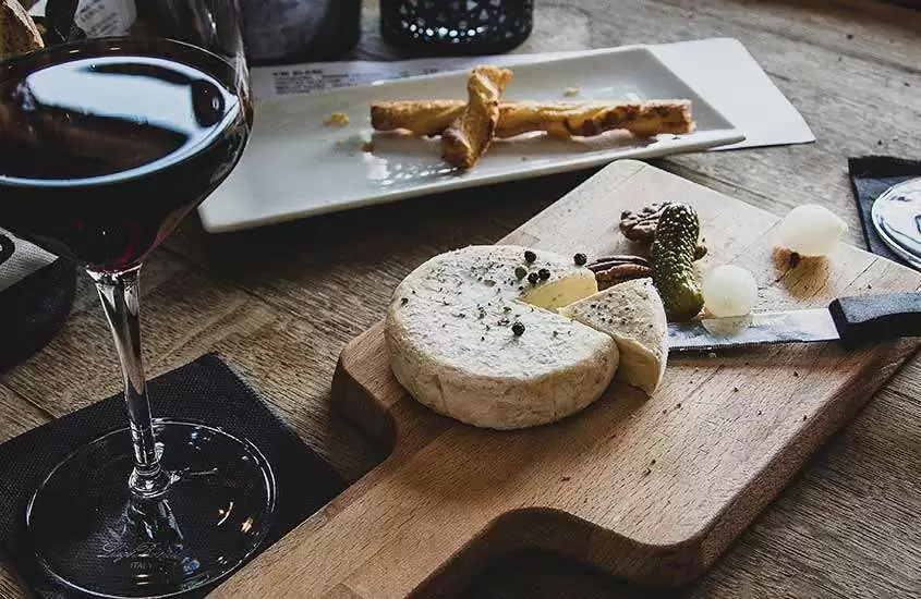 taça de vidro com vidro tinto e pedaço de queijo servido em tábua de madeira em cima de mesa de madeira