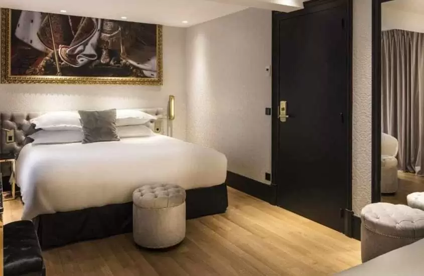 cama de casal e poltrona em suíte de hotel com paredes brancas e piso parquet