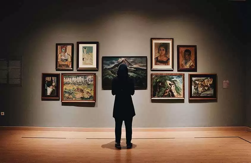 pessoa vestida com roupas pretas, de costas, observando quadros em exposição em um museu.