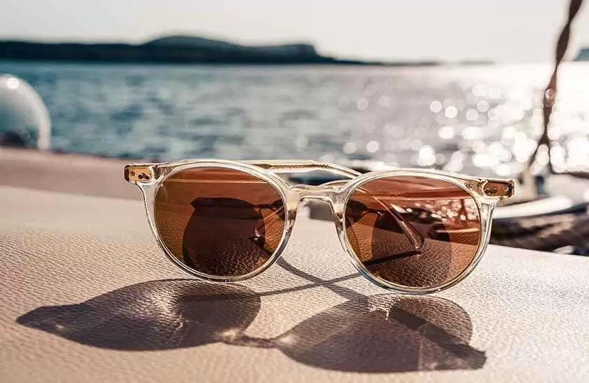Óculos de sol sobre um pano em primeiro plano, com o mar desfocado ao fundo, em uma ensolarada praia de Ibiza.