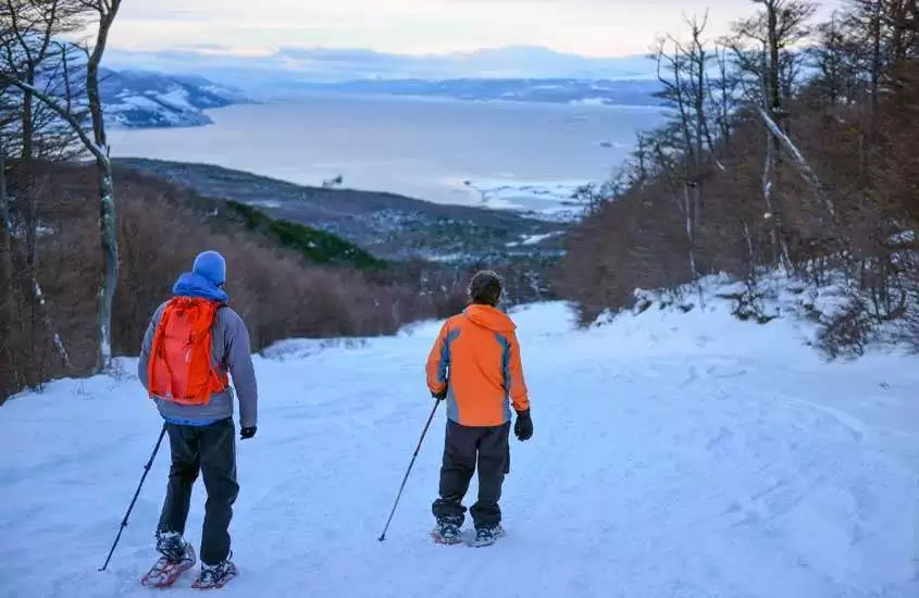 Durante o dia, duas pessoas de costas caminhando em montanha coberta de neve com vista panorâmica de Ushuaia
