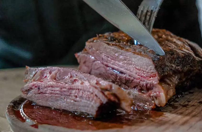 Carne de churrasco em tábua sendo cortada com garfo e faca