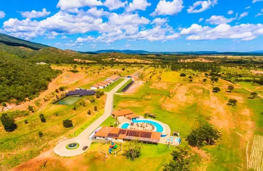 Durante dia de sol, vista aérea panorâmica de um hotel fazenda no Sul de Minas com construções, piscina, lago artificial e muita área verde em volta