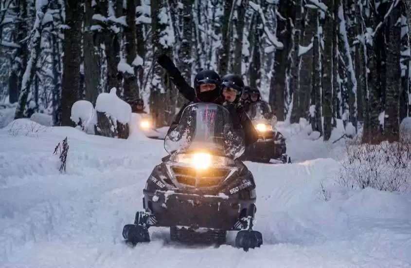 Durante a noite, turistas em motos de neve com farol acesso passeando em bosque coberto de neve